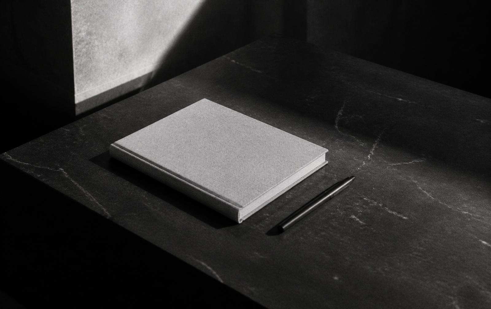 Caderno de linho e caneta sobre mesa de pedra escura sob luz rasante, em preto e branco